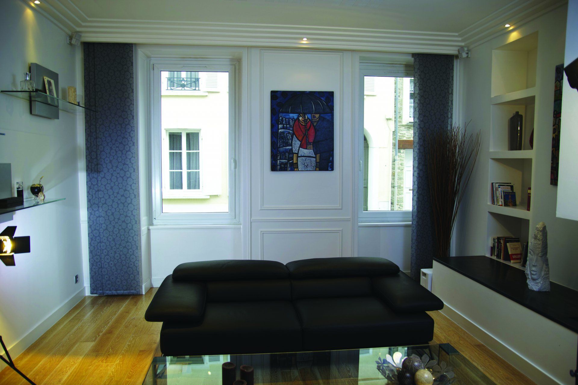 Salon moderne avec canapé noir, table basse en verre et murs blancs ornés de tableaux encadrés.