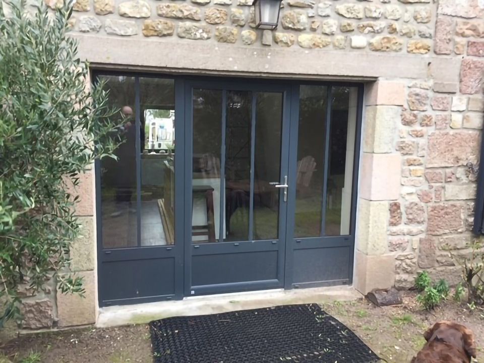 Porte d'entrée double vitrée bleu foncé dans un bâtiment en pierre, avec un paillasson noir et des plantes à proximité