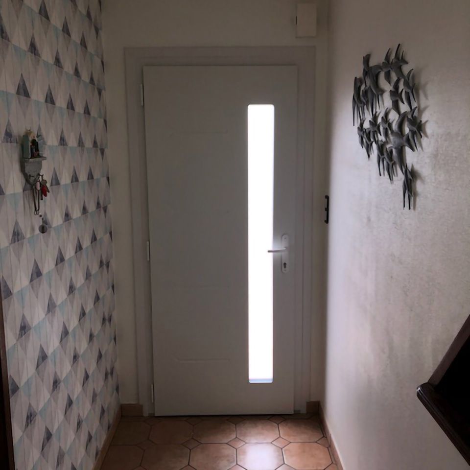 Couloir étroit avec une porte d'entrée blanche, une fenêtre verticale étroite, du papier peint à motifs et une décoration murale.