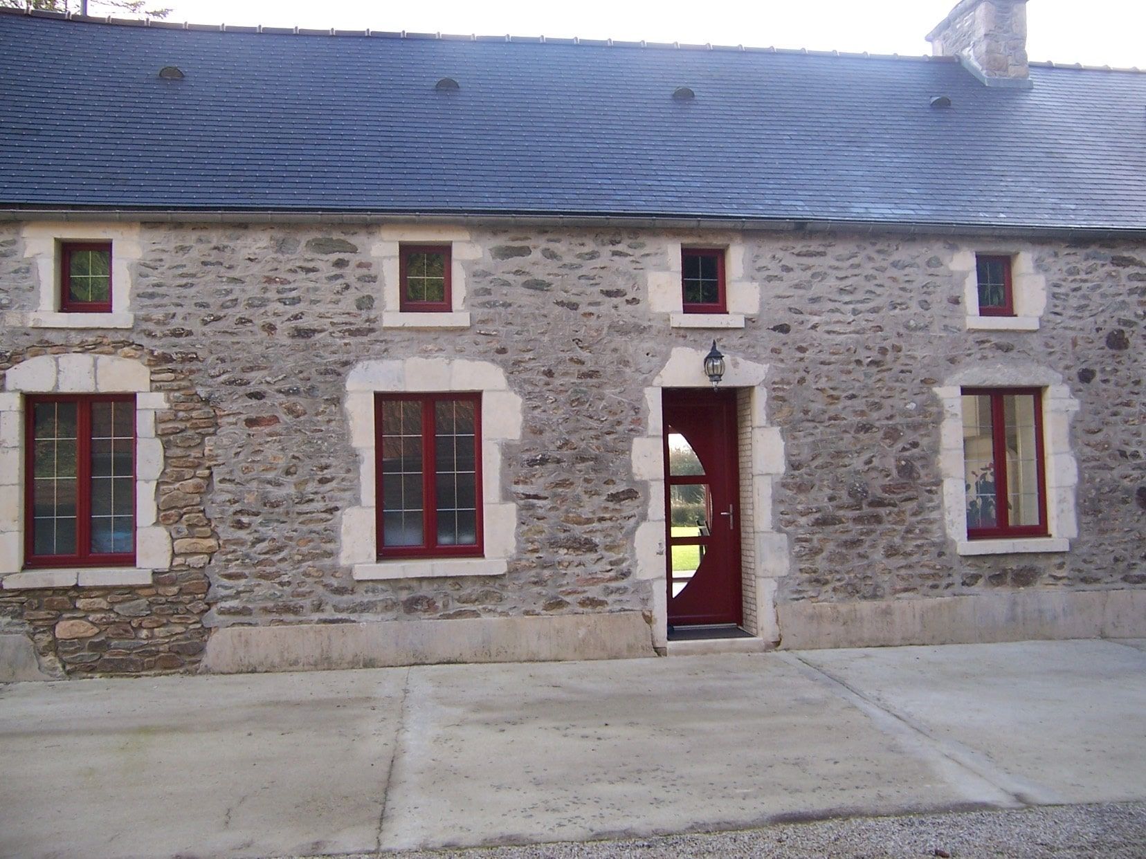 Façade de maison en pierre avec fenêtres à encadrement rouge et porte centrale sous un toit bleu-gris