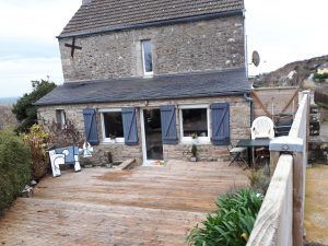 Maison en pierre avec terrasse en bois, fenêtres à encadrement bleu et coin salon extérieur donnant sur une colline