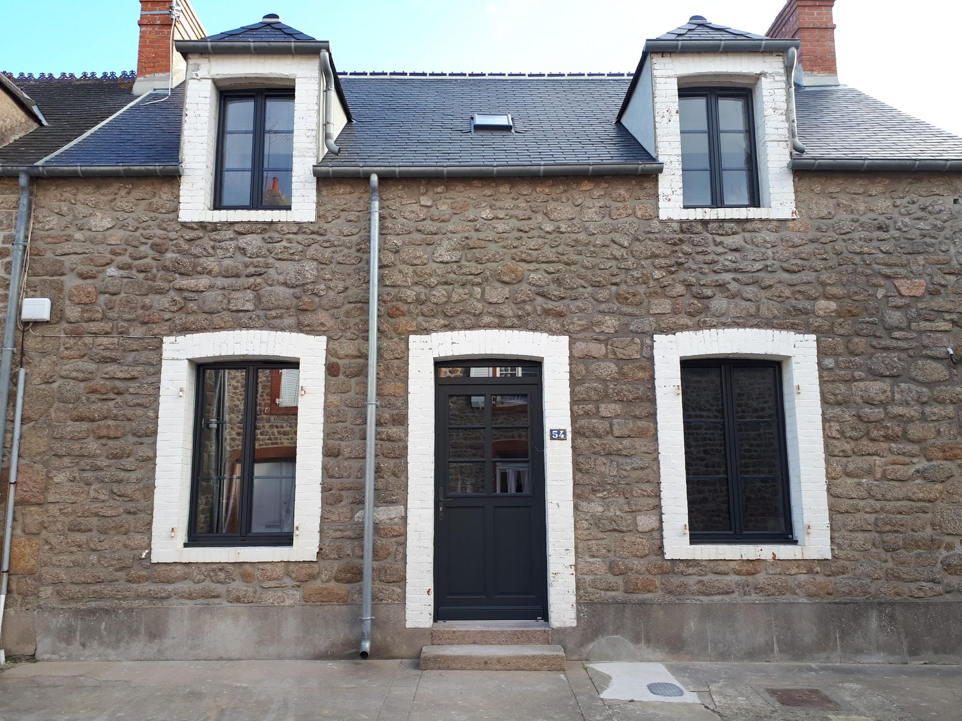 Façade de maison en pierre avec porte bleu foncé et fenêtres à encadrement blanc sous un toit en ardoise