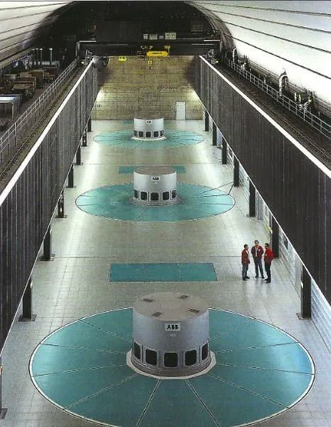 Intérieur d'une centrale hydroélectrique avec trois turbines sur des bases circulaires de couleur turquoise. Des personnes se tiennent au centre.