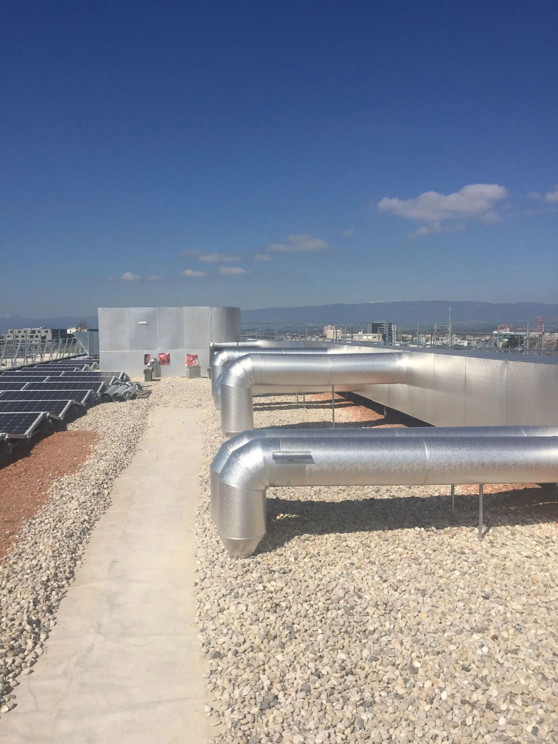 Vue du toit : tuyaux et équipements sur un toit recouvert de gravier sous un ciel bleu.