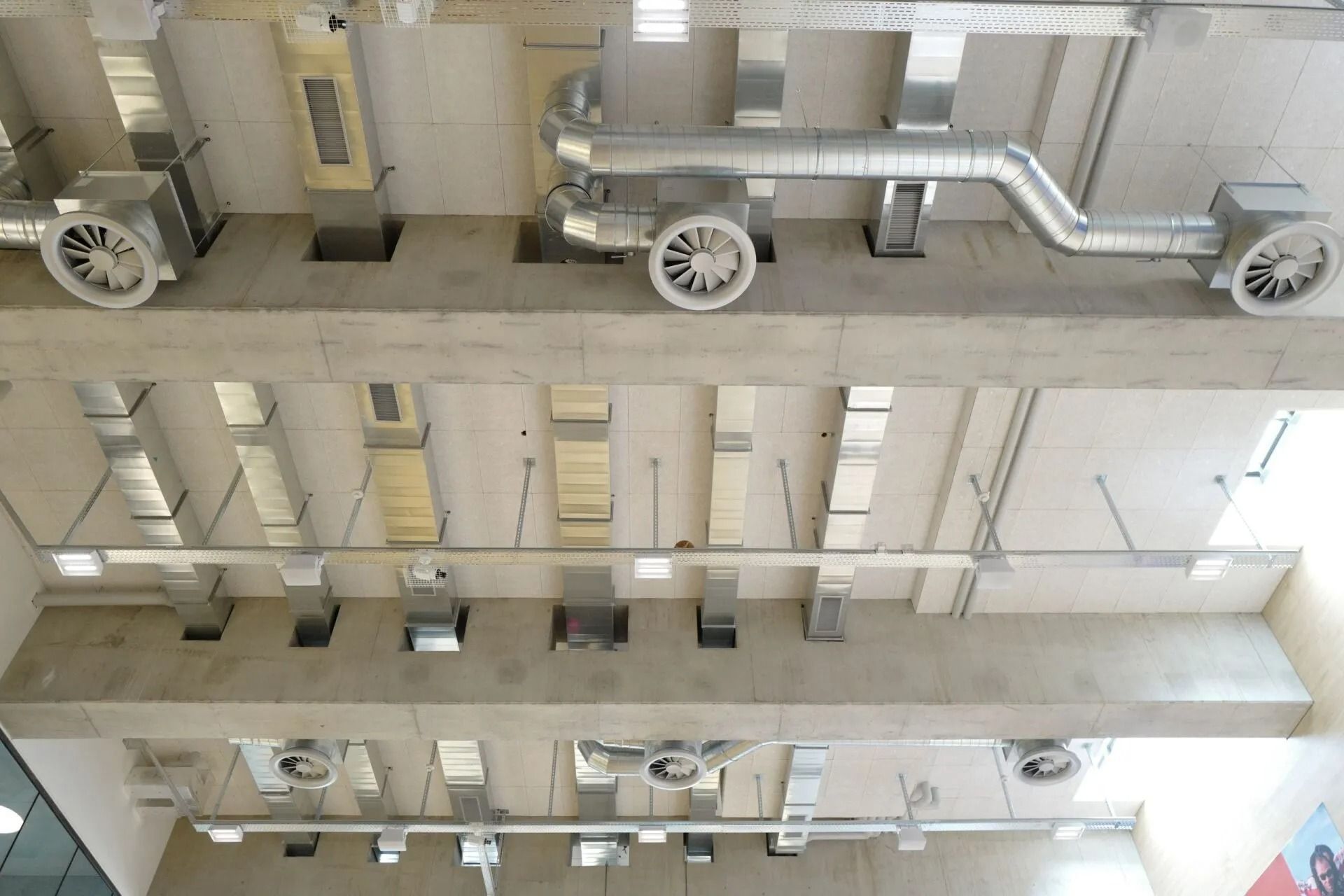 Vue du plafond avec conduits de ventilation, ventilateurs et luminaires dans un bâtiment moderne.