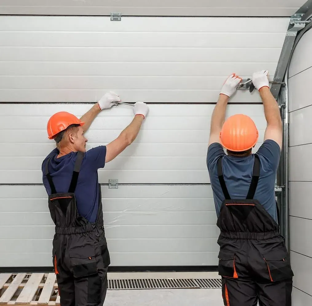 Dos trabajadores con cascos y monos de color naranja reparan una puerta de garaje.
