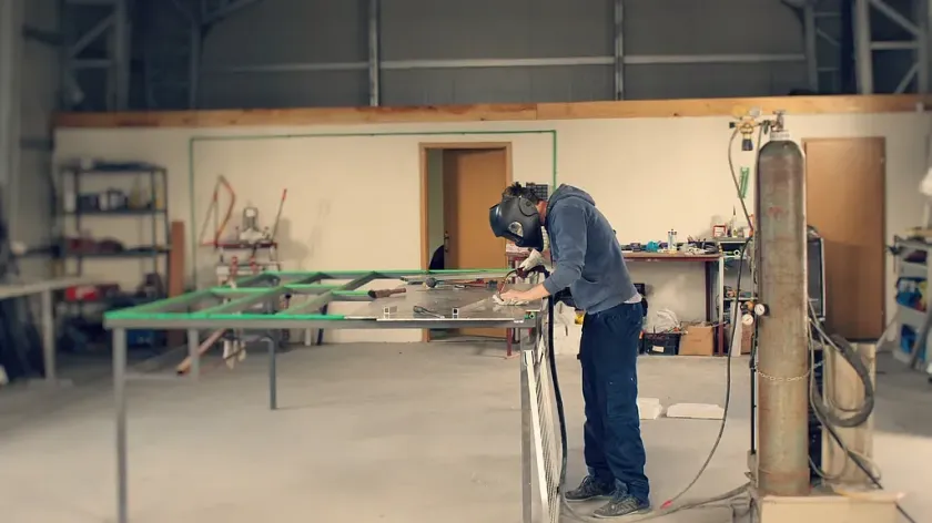 Persona soldando una estructura metálica en un taller. Lleva casco y equipo de protección.