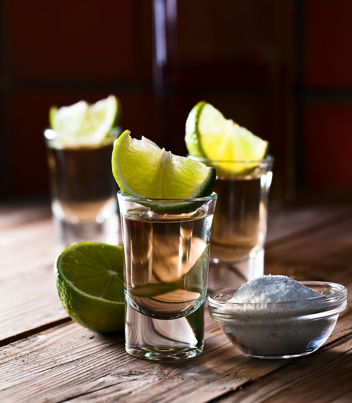 Tres vasos de chupito llenos de tequila y rodajas de lima sobre una mesa de madera.
