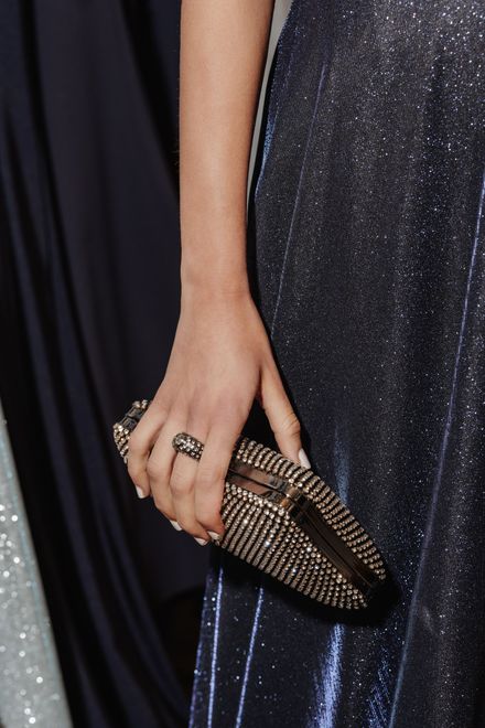 Eine Frau hält eine Clutch mit Perlen und trägt einen Ring. Im Hintergrund ist ein dunkelblaues, glitzerndes Kleid zu sehen.