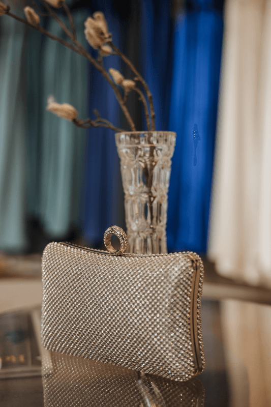 Funkelnde goldene Clutch neben einer Kristallvase mit Zweigen, vor bunten drapierten Stoffen.