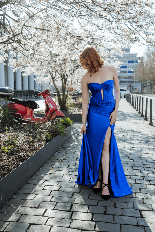 Frau in blauem Abendkleid hält Saum und posiert neben rotem Motorroller und blühendem Baum.