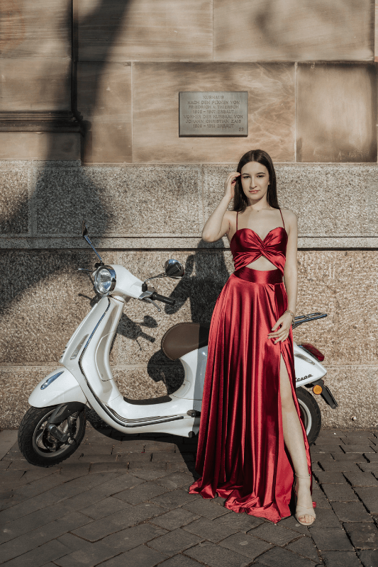 Frau in rotem Kleid posiert neben einem weißen Motorroller vor einem Steingebäude.