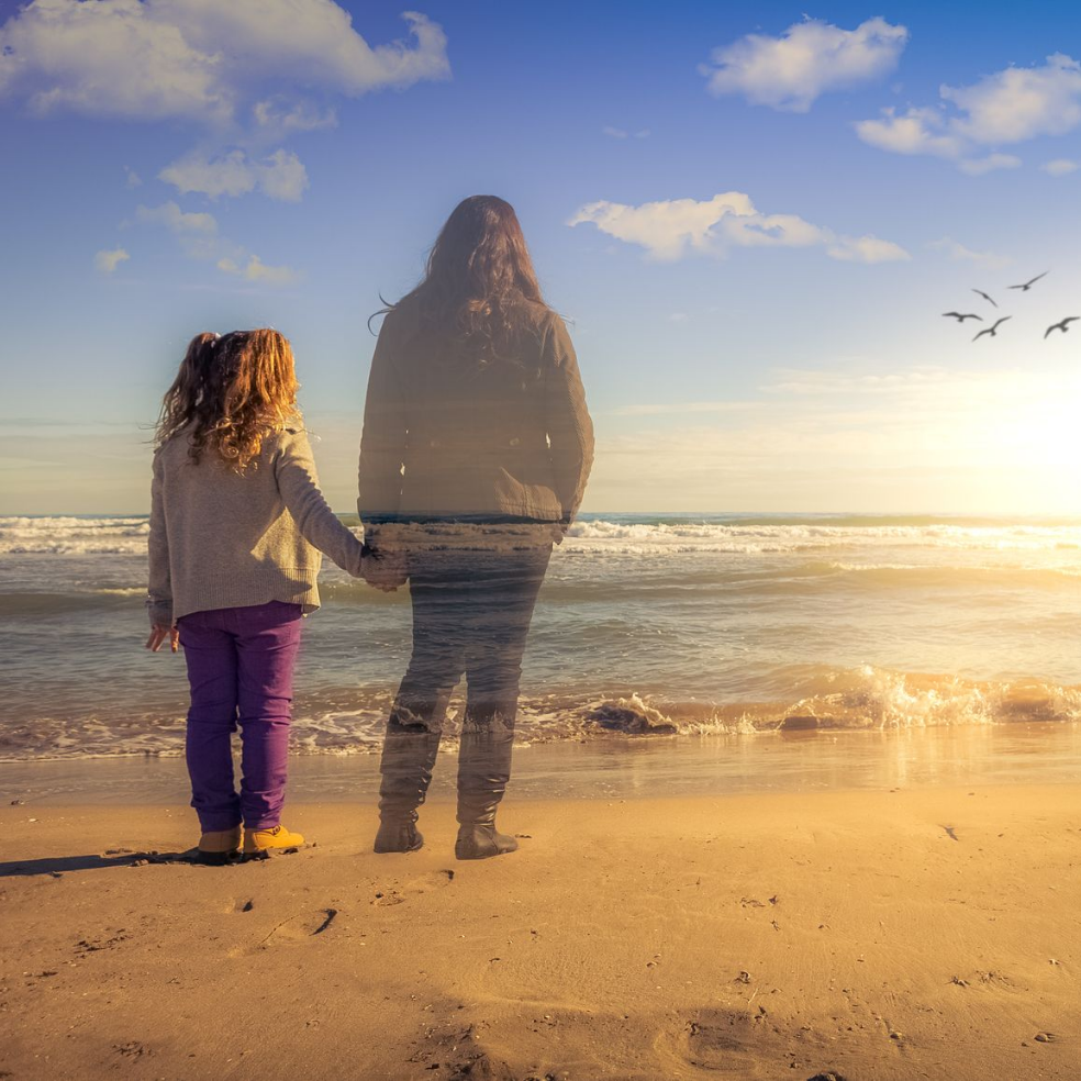 Un enfant et une personne en transparence se tenant la main, debout sur la plage, regardant l'océan.