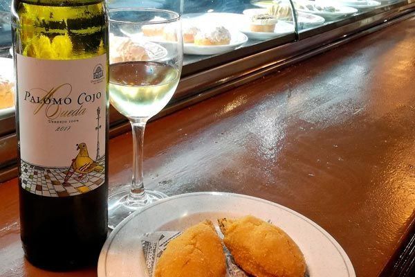 Una botella de vino, una copa de vino y dos pasteles sobre la barra de un bar, junto a una vitrina.