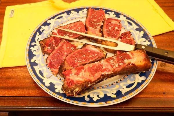 Fileteado de lonchas de carne a la parrilla con pinzas en un plato azul estampado, colocado sobre una mesa de madera con una servilleta amarilla.
