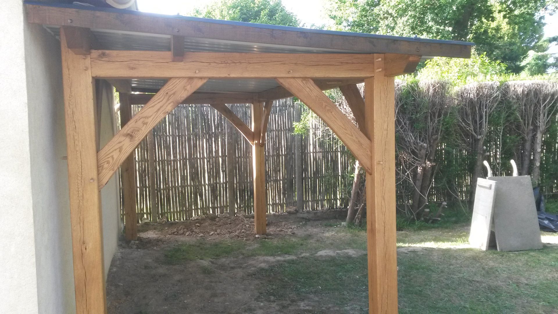Dans une cour, une pergola en bois est adossée à un mur blanc, avec des poteaux de soutien et des entretoises d'angle diagonales.