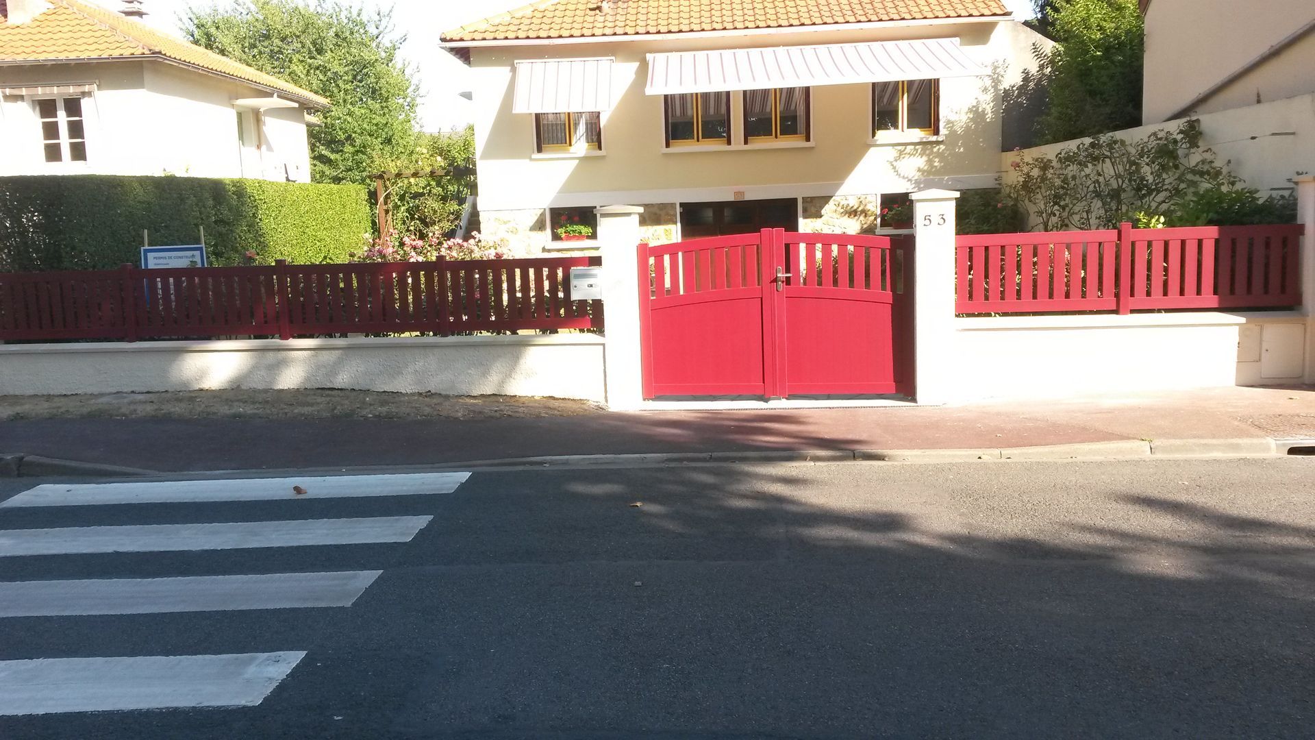 Une maison à la façade beige, à la clôture rouge et au toit de tuiles brunes, vue de la rue avec un passage piéton devant.