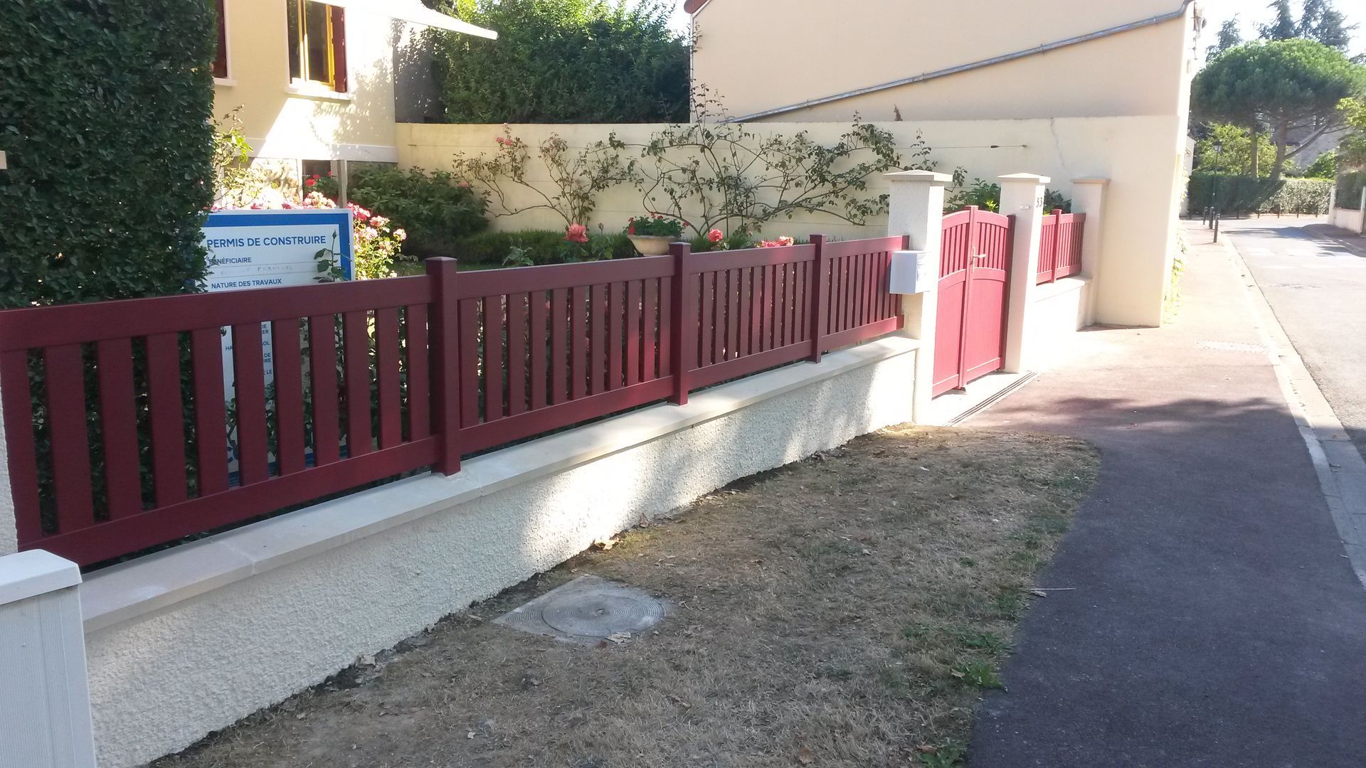 Par une journée ensoleillée, une clôture en bois bordeaux surplombe un muret en pierre blanche devant une maison d'habitation.