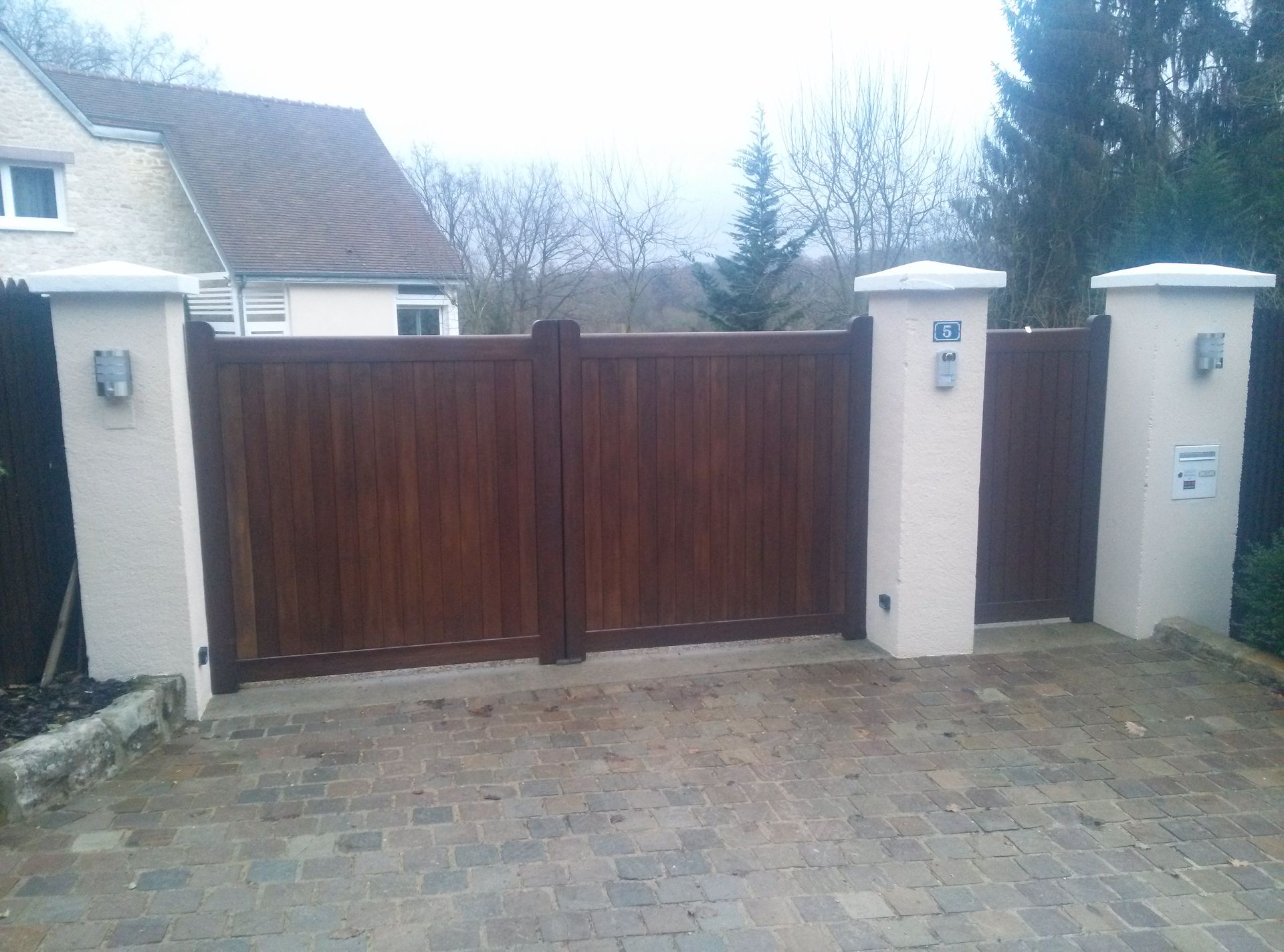 Un large portail double en bois brun foncé, encadré par trois piliers en pierre couleur crème, donne accès à l'allée d'une maison.