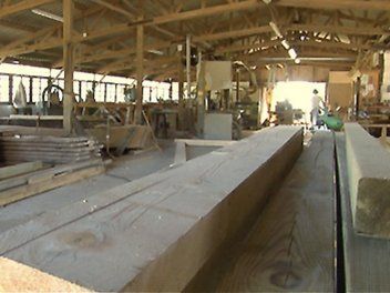 Intérieur d'un atelier de menuiserie avec de grosses poutres en bois empilées au premier plan et des machines à l'arrière-plan.