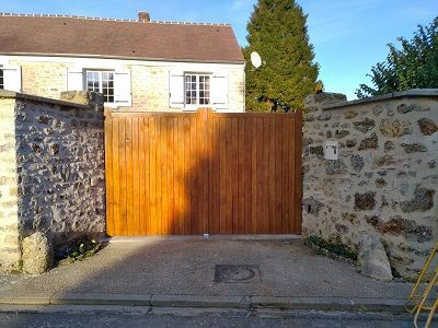Un portail en bois, encadré par deux murs de pierre, se dresse devant une maison sous un ciel d'un bleu limpide.