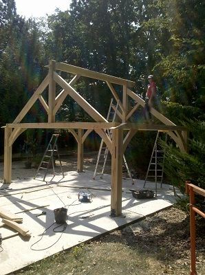 Un ouvrier se tient debout sur la charpente en bois partiellement construite d'une structure extérieure, soutenue par trois échelles.