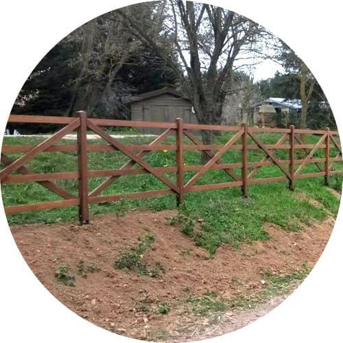 Une clôture en bois à poteaux et traverses avec des entretoises diagonales, installée dans une cour herbeuse près d'arbres et d'un abri de jardin.