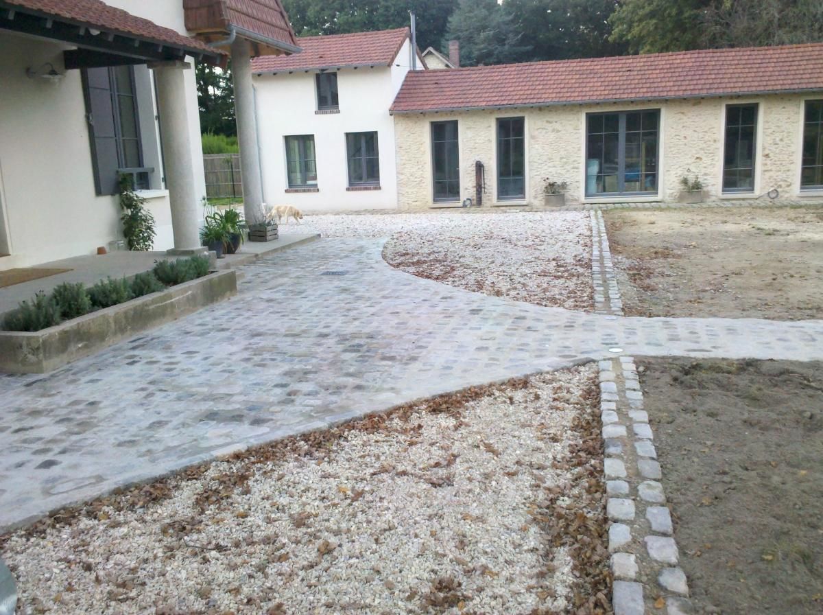 Un chemin pavé de pierres mène à une maison blanche et à un bâtiment en pierre adjacent, sous un toit de tuiles rouges, dans un cadre de jardin.