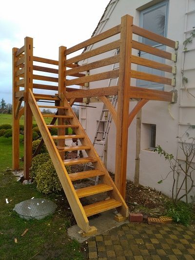 Une terrasse et un escalier en bois sont installés contre le mur extérieur blanc d'une maison, dans un jardin gazonné.