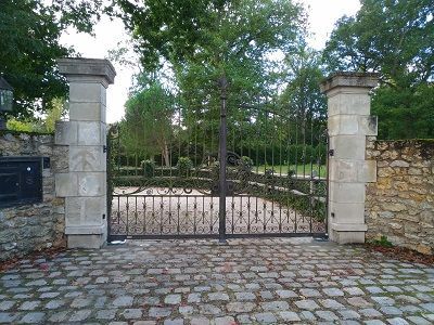 Des grilles en fer forgé flanquées de piliers en pierre et une allée pavée, le tout encadré d'arbres et de murs en pierre.