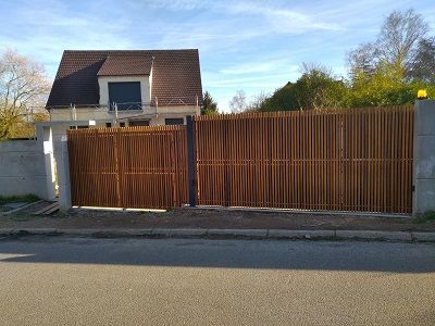 Une maison vue de la rue, derrière une clôture et un portail modernes en lattes de bois.