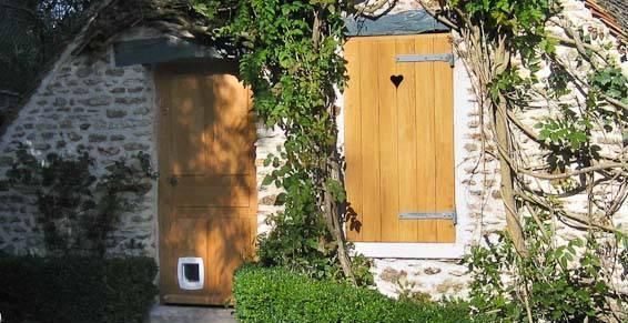 Une maison en pierre avec des portes en bois, une chatière et un volet découpé en forme de cœur, entourée de plantes grimpantes.