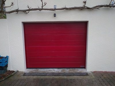 Une porte de garage rouge à panneaux horizontaux encastrée dans un mur extérieur de couleur claire, au-dessus d'une allée pavée.