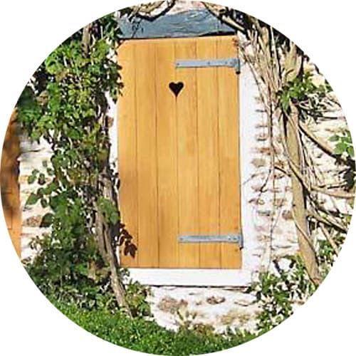 Une porte rustique en bois avec une découpe en forme de cœur, encastrée dans un mur de pierre et entourée de vignes grimpantes et de feuillage envahissants.
