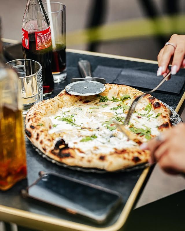 On découpe une pizza sur une table, avec un soda, un verre d'eau et un téléphone.