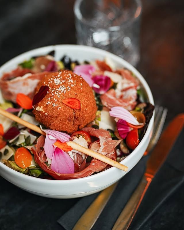 Saladier garni de fritures, de prosciutto, de pétales de fleurs et de couverts dorés sur un fond sombre.