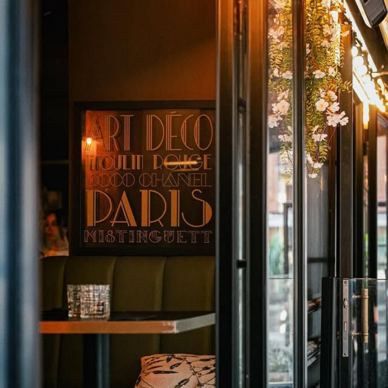 Affiche Art déco dans un café parisien à l'éclairage tamisé, avec du feuillage et une porte ouverte.