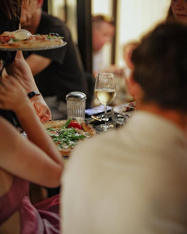 Des personnes dînent dans un restaurant, des pizzas et du vin sont posés sur la table, un serveur tient une pizza.