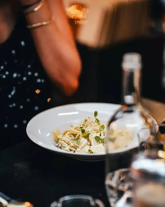 Assiette de pâtes garnie ; personne en arrière-plan ; bouteille au premier plan.