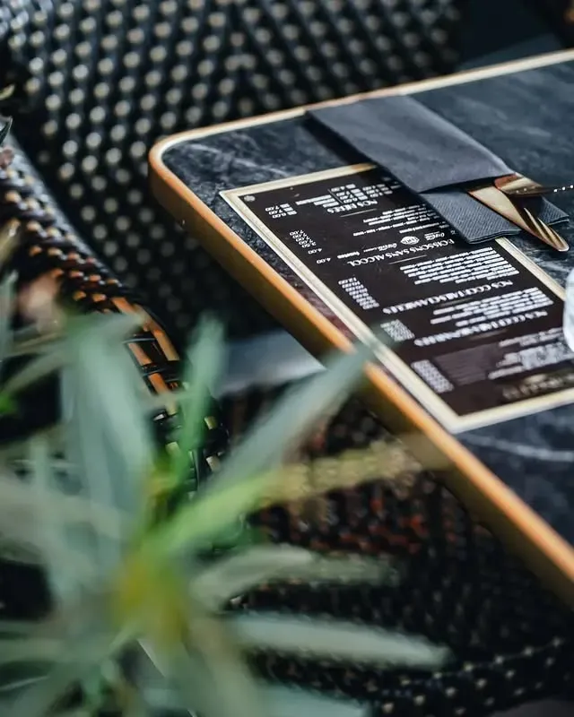 Table dressée avec un set de table noir, un menu, des couverts et une plante au premier plan.