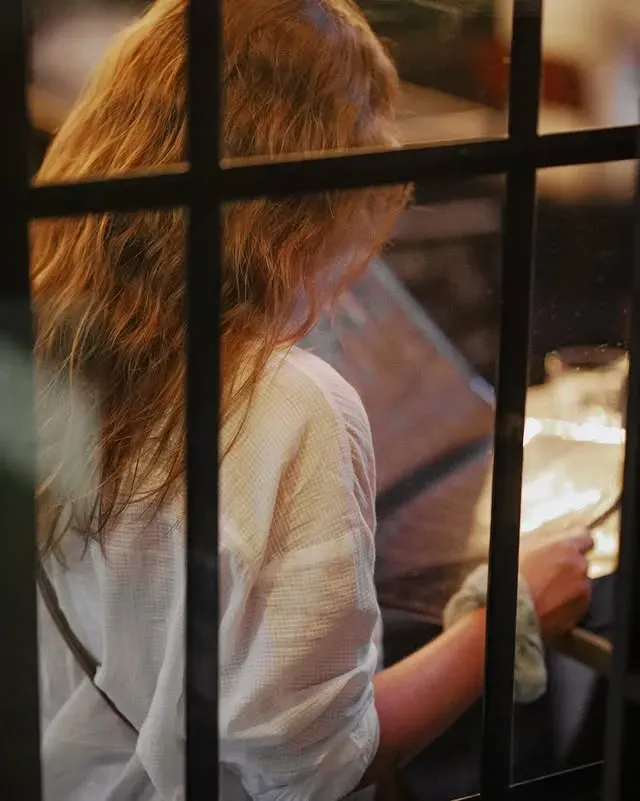 Une femme aux cheveux ondulés mange derrière une fenêtre.