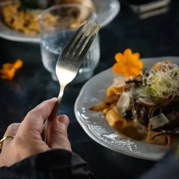 Une main tenant une fourchette avec des pâtes dans une assiette, un verre d'eau et une garniture florale sur une surface sombre.