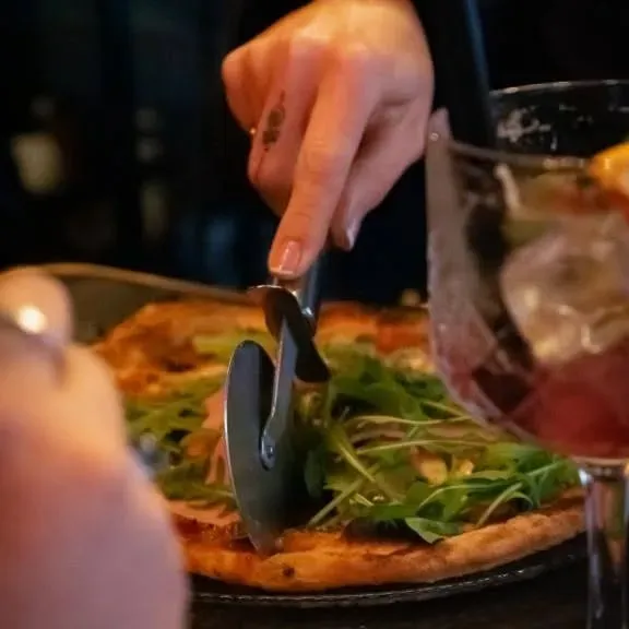 Gros plan d'une personne coupant une pizza avec une roulette. Roquette sur la pizza. Verre de cocktail rouge à droite.