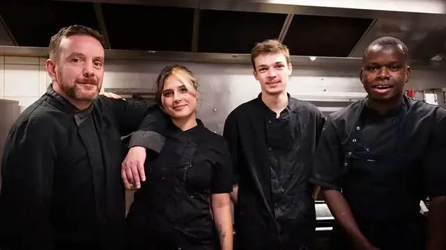 Quatre chefs en uniforme noir posent dans une cuisine professionnelle.