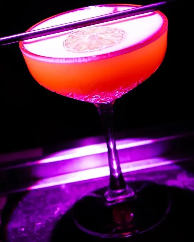 Cocktail servi dans un verre à pied, boisson à l'orange, mousse blanche surmontée d'une tranche d'agrume et d'une paille en métal, lumière violette.