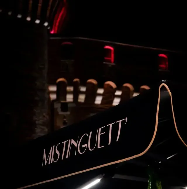 Enseigne de MISTINGUETT' sur fond sombre. Éléments architecturaux et accents rouges en arrière-plan flou.