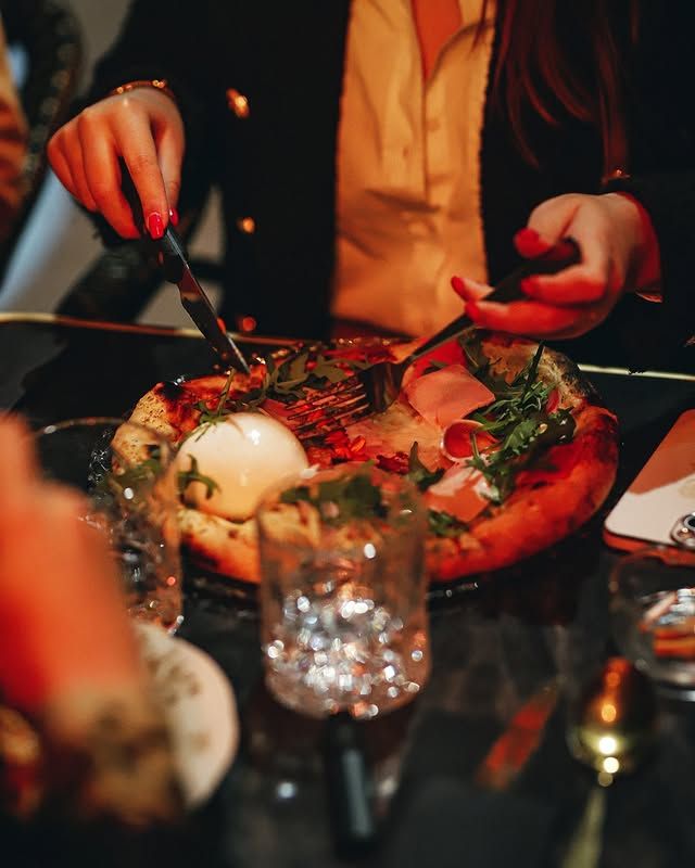 Une personne mange une pizza garnie d'œufs et de légumes verts à l'aide d'un couteau et d'une fourchette, attablée à une table.