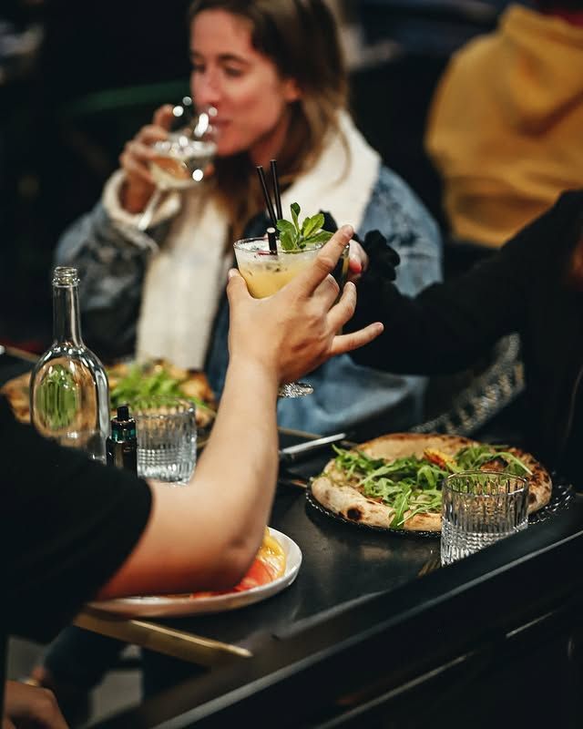 Des personnes attablées dans un restaurant, l'une d'elles sirotant un cocktail. Des pizzas et des verres d'eau sont posés sur une table sombre.