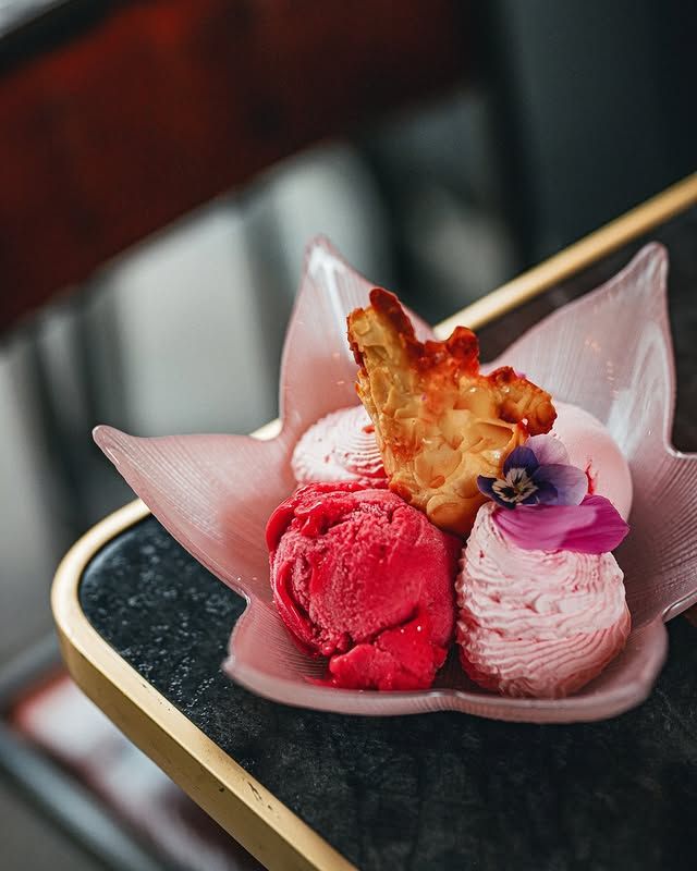 Dessert aux teintes roses composé de boules de glace, d'un biscuit et d'une fleur, présenté dans un plat décoratif en forme de feuille.