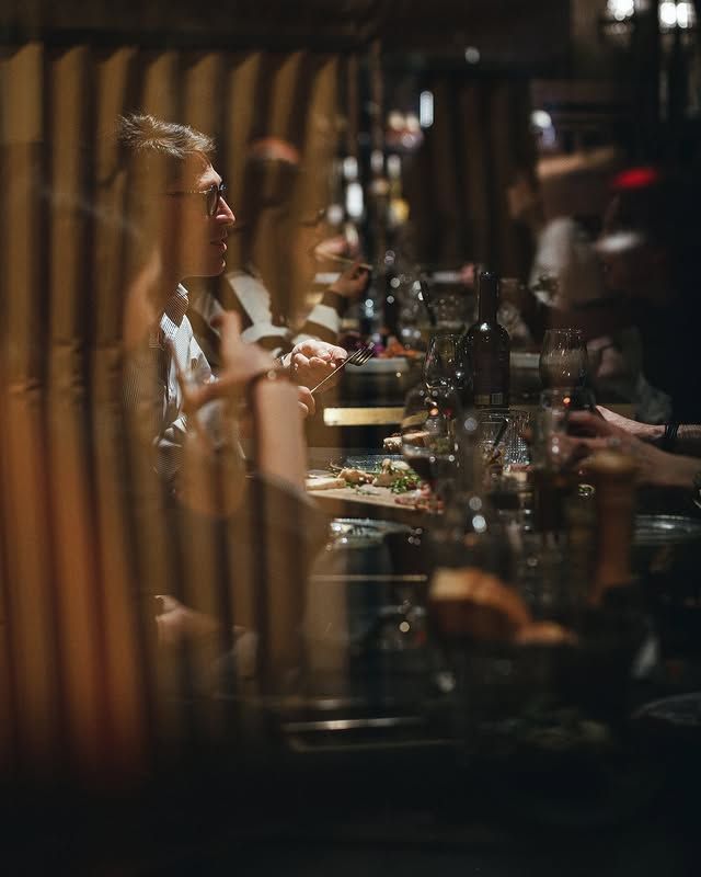 Des clients attablés dans un restaurant, aperçus à travers des lattes de bois verticales. Verres et plats sur la table. Éclairage tamisé.